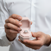 A close-up of a person in a white dress shirt holding an open light pink jewelry box that displays a silver-toned ring with a square-cut clear gemstone.