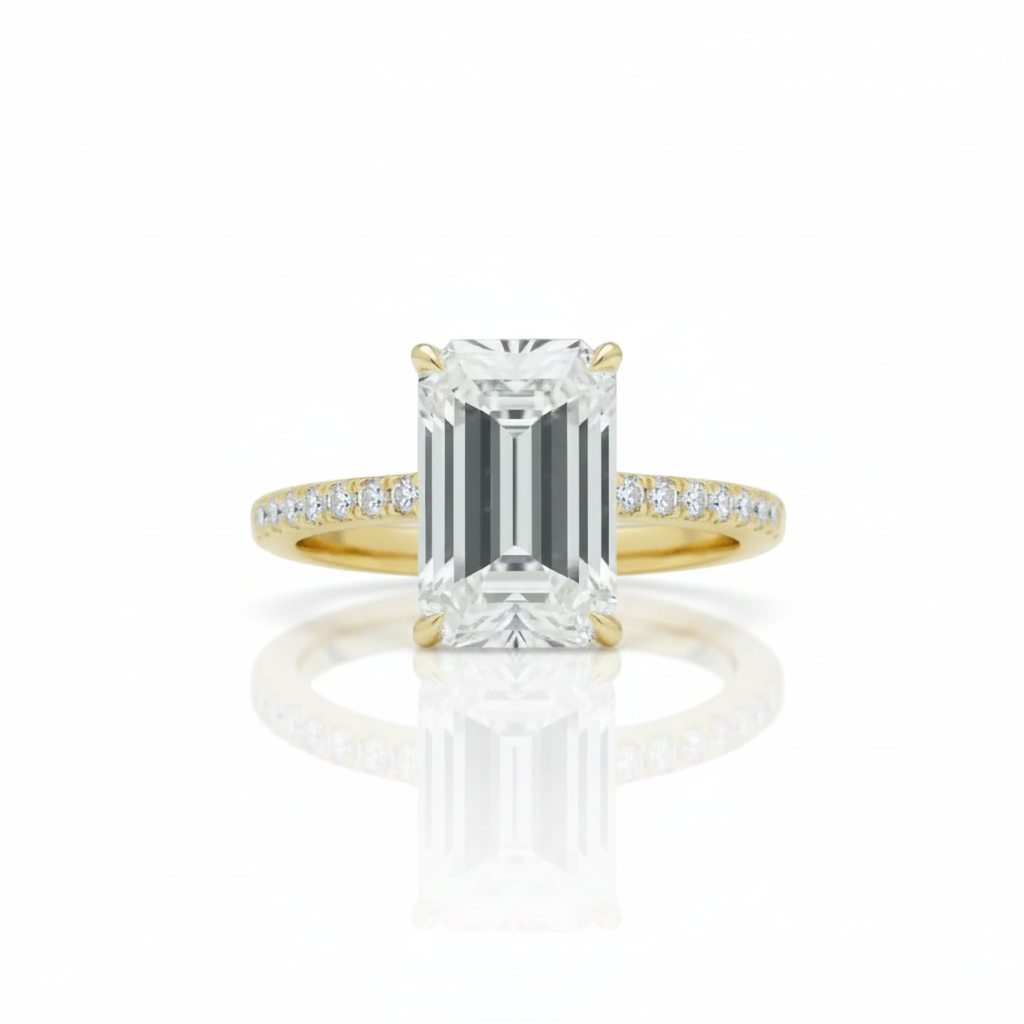 An emerald-cut diamond engagement ring with a yellow gold band accented by small pavé diamonds, shown on a clean white background with a soft reflection below.