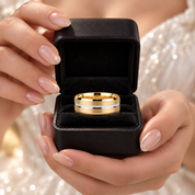 A gold wedding band with a silver inlay displayed inside an open black presentation box, held by hands with manicured nails against a sparkling white dress background.