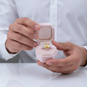 Person holding a pink jewelry box with a diamond ring inside, wearing a white shirt.
