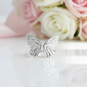 Silver butterfly rings on a reflective surface with pink and white roses in the background