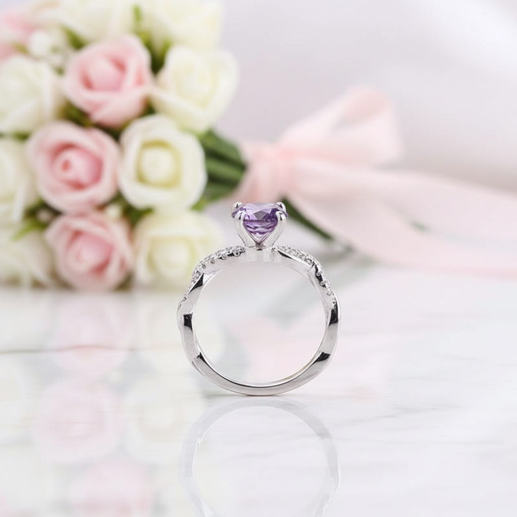 A silver ring with a heart-shaped purple gemstone and small clear stones along the band, displayed on a reflective white surface with pink and white roses in the background.