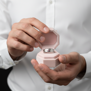 A close-up of a person in a white button-up shirt holding an open light pink octagonal ring box, with a silver ring displayed inside.