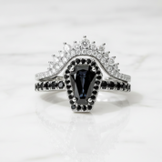 A silver-tone ring set featuring a central marquise-cut dark blue gemstone surrounded by small black stones, with a split band adorned with clear pavé stones and a matching crown-like top, photographed on a light marble surface.