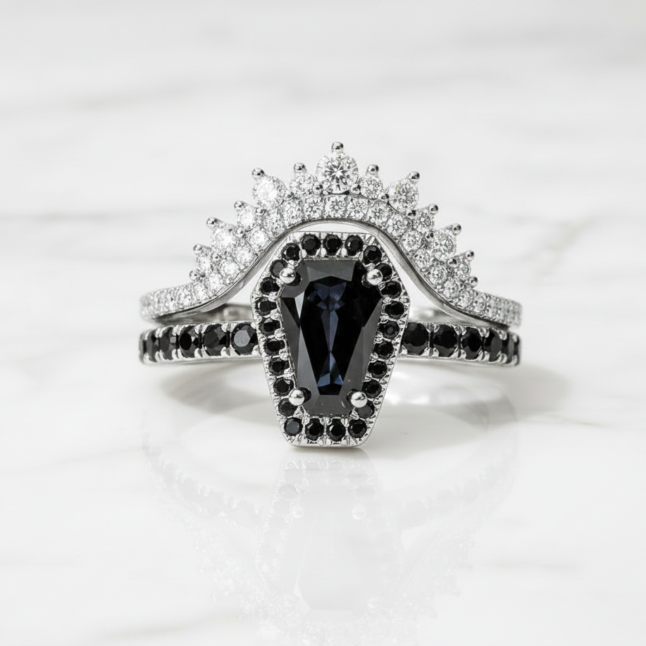 A silver-tone ring set featuring a central marquise-cut dark blue gemstone surrounded by small black stones, with a split band adorned with clear pavé stones and a matching crown-like top, photographed on a light marble surface.