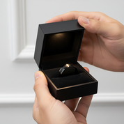 Person holding a black ring box with a ring inside against a neutral background