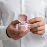 Person holding a pink jewelry box with a diamond ring inside against a white background