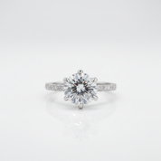 A classic solitaire engagement ring featuring a round brilliant-cut center stone in a six-prong setting on a polished silver-colored band with small pavé accents, shown on a clean white reflective surface.