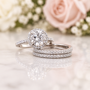 A silver-toned engagement ring with a round diamond halo and matching band, displayed with a coordinating pavé band ring on a light marble surface with soft pink and white flowers blurred in the background.