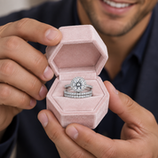A close-up of a person holding an open pink velvet hexagonal ring box, showcasing a silver engagement ring with a round diamond and a matching diamond band.