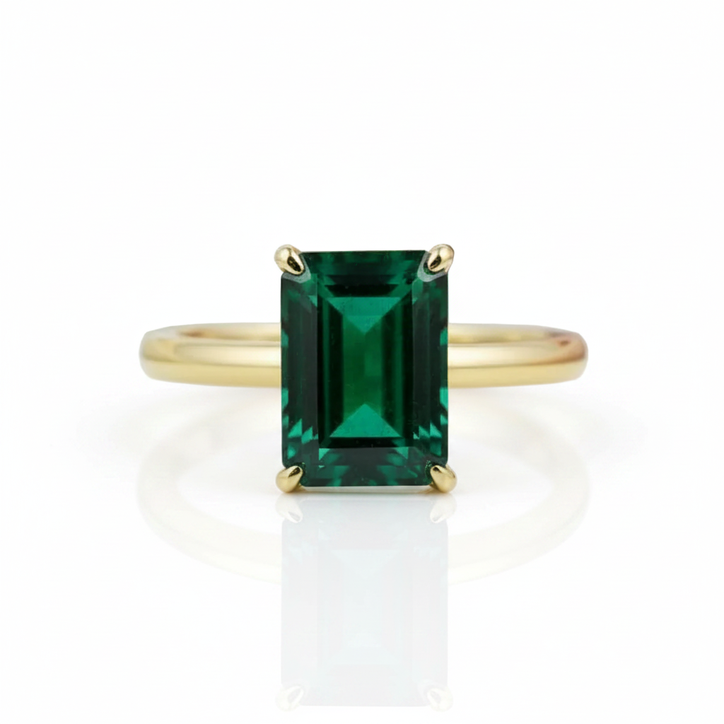An emerald-cut green gemstone ring set in a yellow gold band, shown on a clean white background with a soft reflection below.