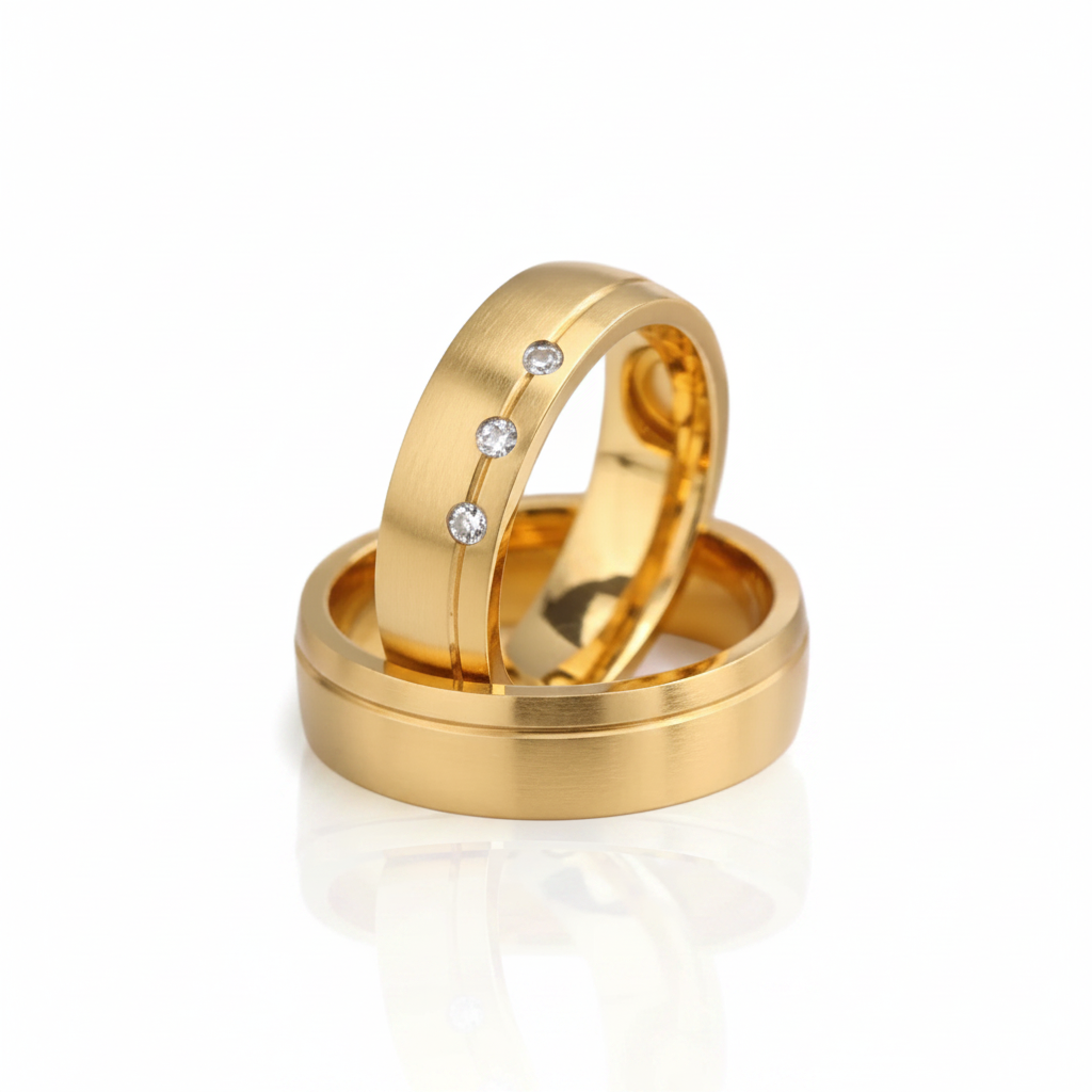 Two gold wedding bands stacked together on a white reflective surface. The top band has three small clear stones set along the side.