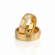 Two gold wedding bands stacked together on a white reflective surface. The top band has three small clear stones set along the side.