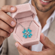 A silver-tone flower pendant necklace with turquoise inlay is displayed in an open pink velvet jewelry box, held by a person wearing a white collared shirt.