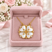 A gold four-petal flower pendant necklace with white iridescent mother-of-pearl inlays and small clear stones, displayed inside an open pink jewelry box on a marble surface with soft pink ribbons and flowers in the background.