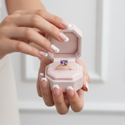 Hand holding a pink jewelry box with a ring featuring an amethyst stone.