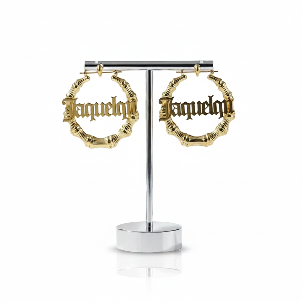 A pair of gold hoop earrings with a bamboo-inspired textured rim and the word 'Jaquelin' in gothic lettering, displayed on a silver T-stand against a white background.