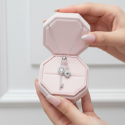 Pink jewelry box with a silver necklace featuring a heart-shaped pendant held by a hand.