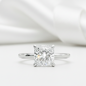 A radiant cut diamond solitaire engagement ring in a four-prong setting on a polished silver band, presented on a soft white background with draped satin fabric.