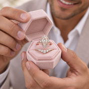 A close-up lifestyle shot of a man holding an open light pink velvet ring box. Inside are three diamond rings: a large pear-cut center stone ring, a smaller marquise-cut ring, and a diamond-accented band.