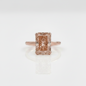An emerald-cut peach diamond ring set on a rose gold band, centered against a clean white background with a soft reflection below.