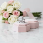 A silver-toned diamond engagement ring with a matching pavé band and a pink velvet ring box, styled on a white marble surface with a bouquet of pink and white roses in the background.