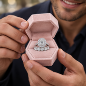 A close-up lifestyle shot of a man holding an open dusty pink velvet ring box containing a silver halo engagement ring and matching band with clear stones.