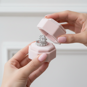 A close-up lifestyle shot of a person's hands holding an open light pink hexagonal ring box, displaying a silver-toned ring set with a large heart-shaped cluster of clear stones and a matching band underneath.