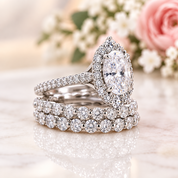 A white gold or platinum engagement ring set with a marquise-cut center stone and halo, displayed on a matching triple-row diamond band set on a polished marble surface with soft pink and white flowers in the background.