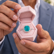 A close-up lifestyle shot of a man holding an open pink velvet hexagonal ring box, showcasing a silver ring with a large cushion-cut turquoise gemstone and small accent diamonds.