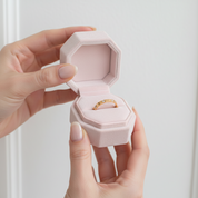 A person's hands holding an open light pink octagonal velvet ring box, which contains a gold band ring with small clear stones, against a soft white background.