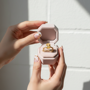 Hand holding a pink jewelry box with a gold ring inside against a neutral background
