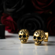 Gold skull-shaped earrings on a reflective surface with a dark floral background
