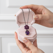 Purple floral necklace in a pink jewelry box held by hands with light nail polish.
