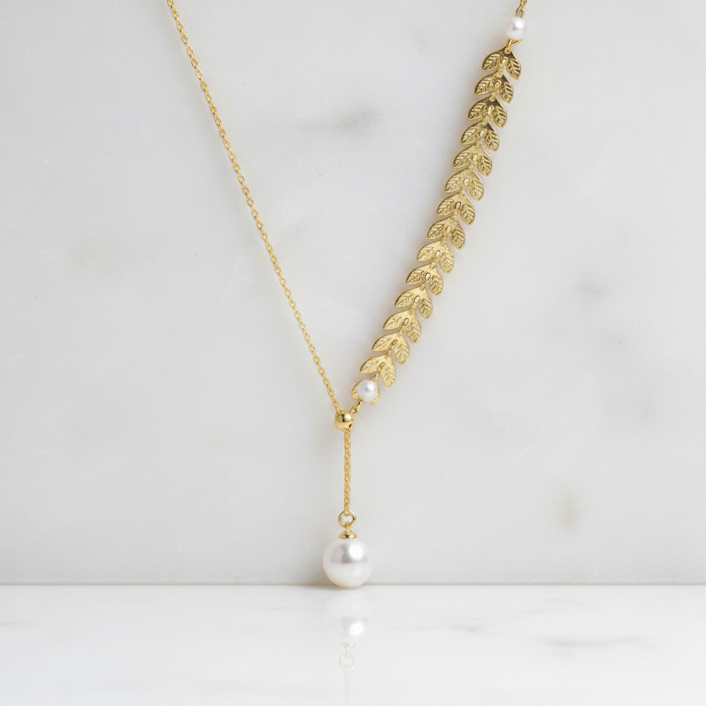 A gold leaf pendant necklace with a single white pearl dangle, displayed on a white marble surface.