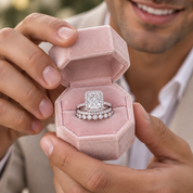 A close-up lifestyle shot of a man holding an open dusty pink velvet ring box. Inside the box are a silver engagement ring with a large square-cut diamond and a matching diamond band.
