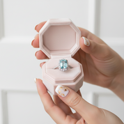 Hand holding a pink hexagonal ring box with an aquamarine ring inside against a white background