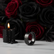 A matte black ring with a small skull-and-crossbones engraving stands on a white marble surface, styled with black wax candles and a background of black and deep red roses.