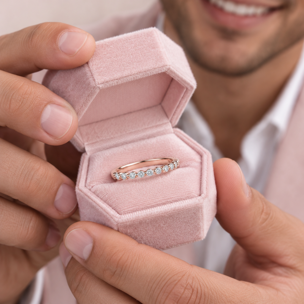 A close-up lifestyle shot of a person holding an open hexagonal pink velvet ring box, showcasing a rose gold band ring with a row of clear gemstones.