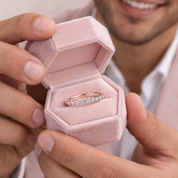 A close-up lifestyle shot of a person holding an open hexagonal pink velvet ring box, showcasing a rose gold band ring with a row of clear gemstones.