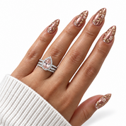 A close-up of a hand with long almond-shaped nails decorated in rose gold and gold floral patterns, wearing a matching rose gold ring with a pear-cut center stone and pavé accents.