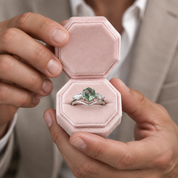 A close-up of a person holding an open pink velvet ring box, displaying a silver ring with a green marquise-cut center stone and small side stones.