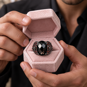 A close-up of a person holding an open pink velvet ring box, showcasing a black oval-cut ring with a textured, beaded silver-toned halo.