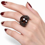 A close-up of a hand wearing a large smoky brown faceted gemstone ring with ornate dark metal detailing, against a clean white background.