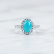 An oval halo ring featuring a vibrant turquoise-colored center stone surrounded by a halo of small clear stones, set on a silver-toned band with pavé accents, resting on a white marble surface.
