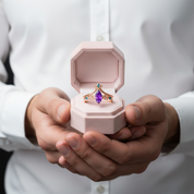 A close-up of a person in a white button-up shirt holding an open light pink jewelry box. The box contains a rose gold ring with a large purple marquise-cut gemstone and small teal accents.