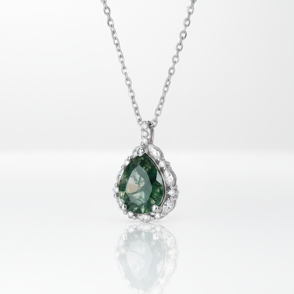 A silver pendant necklace featuring a teardrop-shaped green gemstone surrounded by small clear stones, suspended from a delicate silver chain on a white reflective surface.