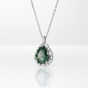 A silver pendant necklace featuring a teardrop-shaped green gemstone surrounded by small clear stones, suspended from a delicate silver chain on a white reflective surface.