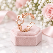 A rose gold open ring with a heart and script letter design, displayed on a pink velvet jewelry box with a soft-focus background of pink roses and white flowers.