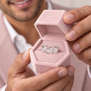 A silver heart-shaped ring with interlocking circular motifs is displayed inside an open pink velvet ring box, held by a person wearing a light pink blazer and white collared shirt.